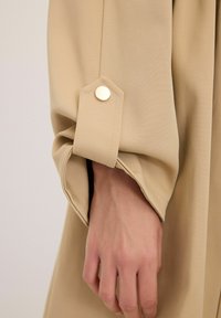 Beige tygärm med en hexagonal manschett och en guldfärgad knapp, som visar en hand med synliga ådror mot en texturerad bakgrund.