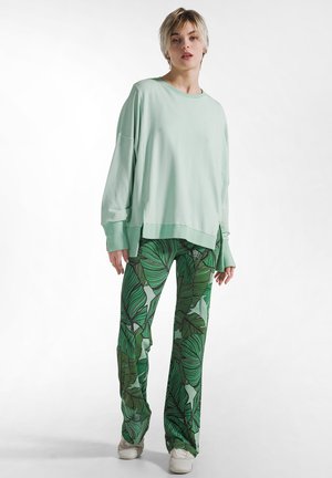 Mátově zelená oversized mikina a kalhoty se vzorem zelených listů, s rozšířenými nohavicemi a volným střihem. Outfit doplňují bílé tenisky.