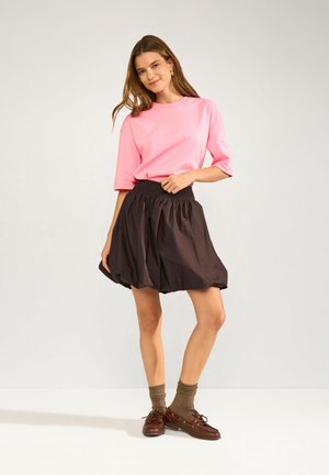 Femme portant un T-shirt rose, une jupe bouffante noire, des chaussettes marron et des chaussures en cuir marron, debout devant un fond clair uni.