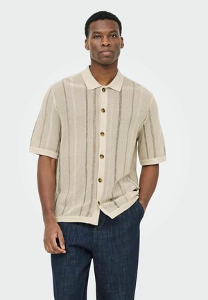 Uomo che indossa una maglietta beige a maniche corte con bottoni in maglia e jeans blu scuro, in piedi con una mano in tasca su sfondo bianco.