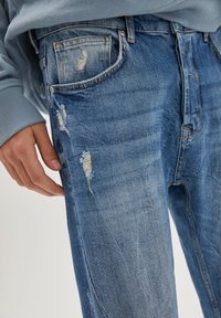 Ingrandimento di una persona che indossa jeans in denim blu strappati con toppe sfilacciate, mostrando la tasca anteriore e la mano poggiata vicino alla coscia.