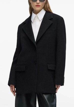 Vrouw met een oversized zwarte blazer, wit overhemd met kraag, zwarte leren broek en ronde donkere zonnebril tegen een witte achtergrond.