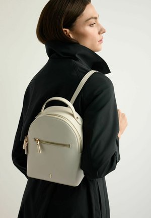 Kobieta w czarnym płaszczu niesie na plecach mały, kremowy skórzany plecak ze złotymi suwakiem, stoi bokiem na jednolitym tle.