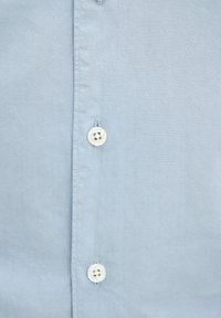 Hellenblaues Hemd mit glatter Textur, ausgestattet mit weißen Knöpfen und geradem Schnitt. Die Knöpfe sind im Vierloch-Stil mit gestickten Details.