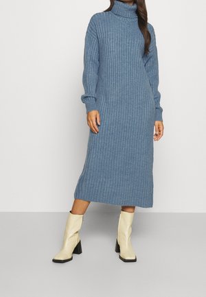 Femme portant une robe midi bleu côtelée à col roulé associée à des bottines à talons crème à bout carré, debout devant un fond uni.