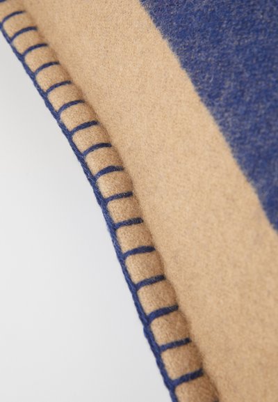 Wollen deken met afwisselende beige en blauwe panelen, met een blauwe gestikte rand voor een gestructureerde afwerking. Zacht en gezellig gevoel.