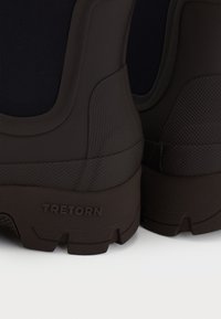 Primer plano de botas de lluvia de goma marrón oscuro con detalles texturizados y el logo de la marca "Tretorn" en el talón.