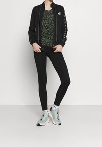 Zwarte zip-up jas met trekkoorden, groene luipaardprint top, zwarte legging en lichtgrijze sport schoenen. Kenmerken logo accenten.