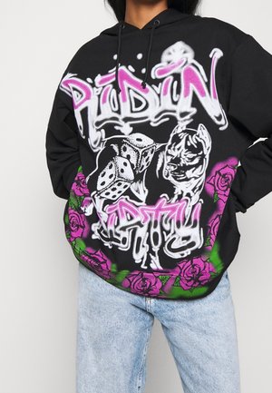 Sweat à capuche noir avec texte en style graffiti "Ridin' Pretty", dés, illustration de chien et roses violettes le long du bas, porté avec un jean bleu clair.