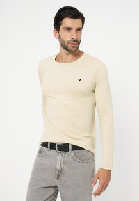 Bărbat cu păr închis la culoare și barbă, purtând o cămașă crem cu mâneci lungi și blugi gri deschis cu curea neagră, stând în fața unui fundal alb simplu.