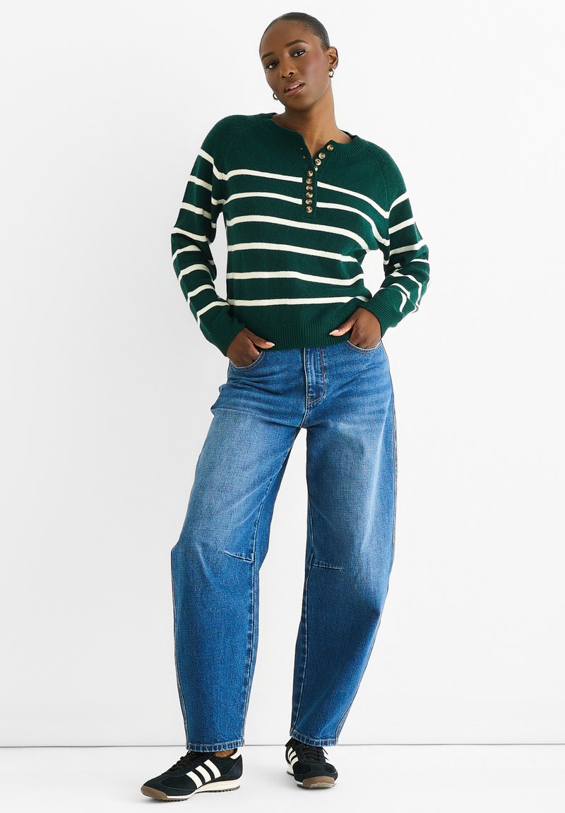 Grün gestreifter Pullover mit Henley-Ausschnitt, versehen mit großen Knöpfen, kombiniert mit hellblauen weit geschnittenen Jeans und schwarzen Sneakers.