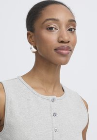 Junge Frau mit dunkler Haut und zurückgebundenem Haar trägt ein ärmelloses, hellgraues, geknöpftes Oberteil und große silberne Creolen.