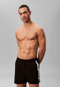 Man zonder shirt met kort haar, draagt zwarte Calvin Klein zwembroek met witte zijstrepen, staand tegen een effen lichtgrijze achtergrond.