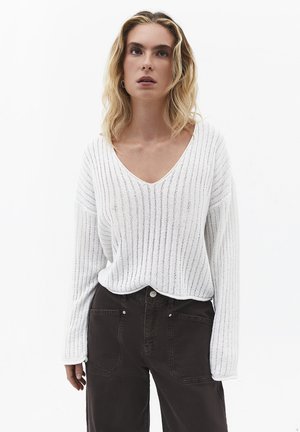 Kvinde med skulderlangt blondt hår iført en hvid strikket V-hals sweater og højtaljede sorte bukser mod en ensartet hvid baggrund.