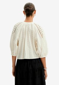 Blusa bianca con maniche a sbuffo, caratterizzata da motivi floreali ricamati e dettagli in pizzo lungo le spalle. Design corto, tessuto leggero.
