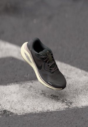 Czarny but do biegania Nike z białą podeszwą unoszący się nad powierzchnią asfaltu z białym oznaczeniem strzałki.