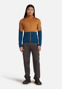 Hombre con cabello largo que lleva una sudadera con cremallera en tonos naranja y azul, pantalones gris oscuro y zapatos de senderismo negros, de pie contra un fondo sencillo.