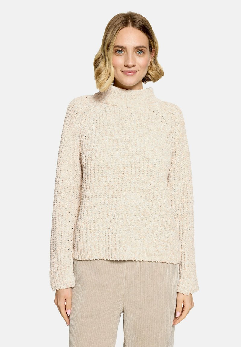 Femme aux cheveux blonds courts portant un pull en laine couleur crème et un pantalon en velours côtelé beige, souriante, mains détendues le long du corps.