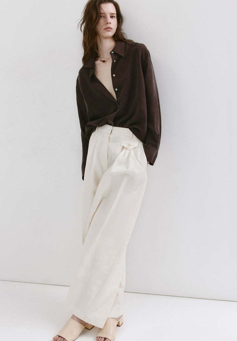 Jeune femme portant une chemise ample marron foncé, un pantalon large couleur crème, et des sandales beiges à talons carrés, debout contre un mur blanc.