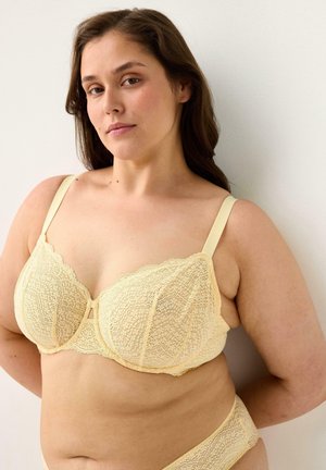 Kvinde med langt brunt hår iført en lysegul blonde-bh og matchende undertøj, stående op ad en hvid væg med hænderne bag ryggen.