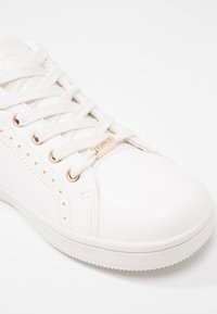 Vit lädersneaker med platta vita snören, metallaccent i guldton och subtila perforeringar längs sidan. Gummisula.