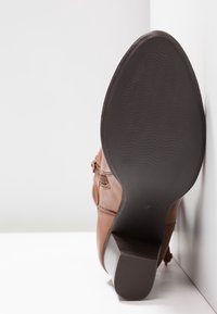 Brown leather ankle boot with a textured black rubber sole, curved heel, and decorative side accents. Shoe positioned on a white background.