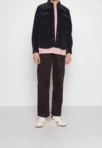 Granatowa koszula sztruksowa na guziki, jasnoróżowa koszulka z okrągłym dekoltem pod spodem, brązowe spodnie sztruksowe oraz białe trampki z czerwonymi detalami.