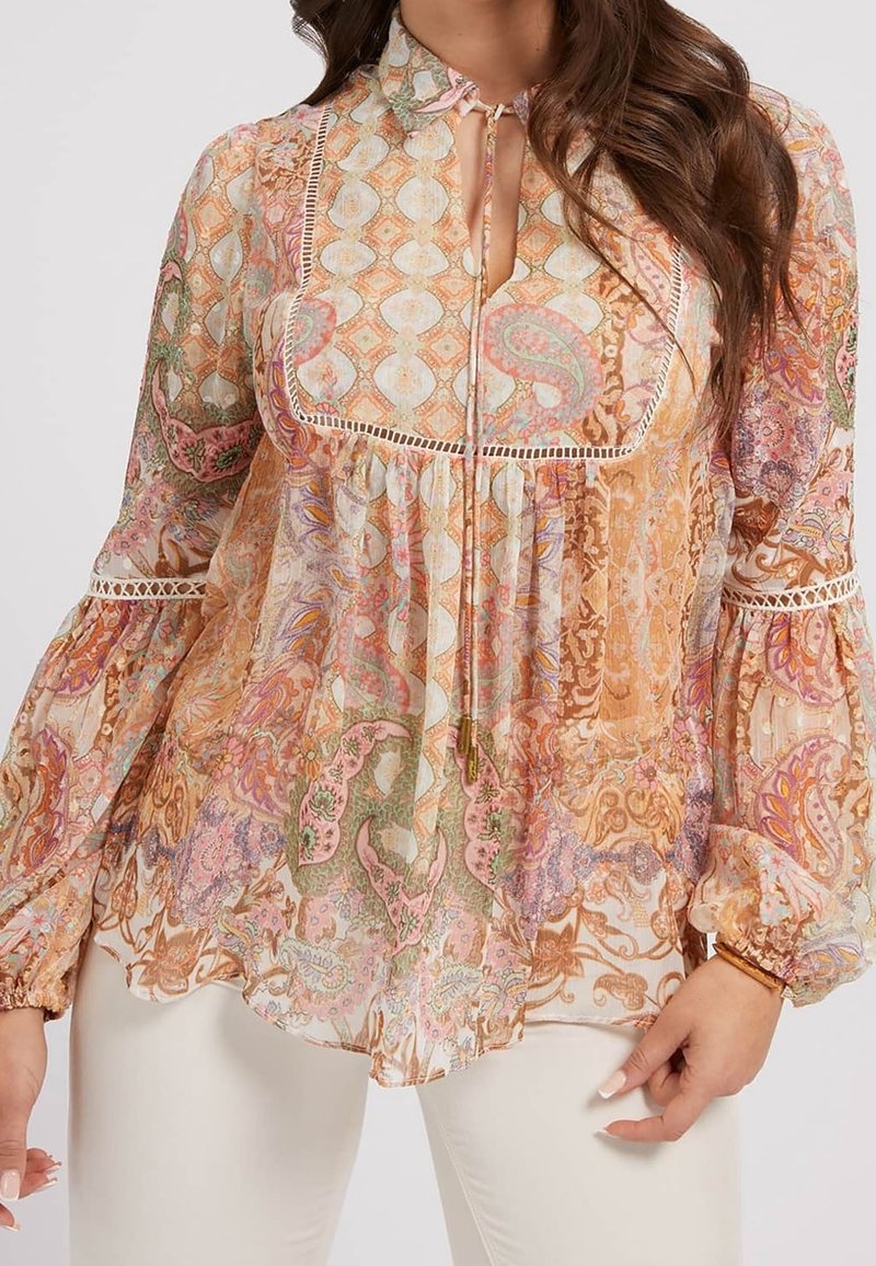 Femme portant un chemisier à manches longues avec des motifs paisley et géométriques dans des tons doux orange, rose, vert et blanc.