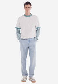 Lässiges Outfit mit einem weißen T-Shirt mit türkisfarbenen Akzenten über einem gestreiften Langarmshirt, hellblauen Jeans und grauen Sneakers mit Gummisohlen.