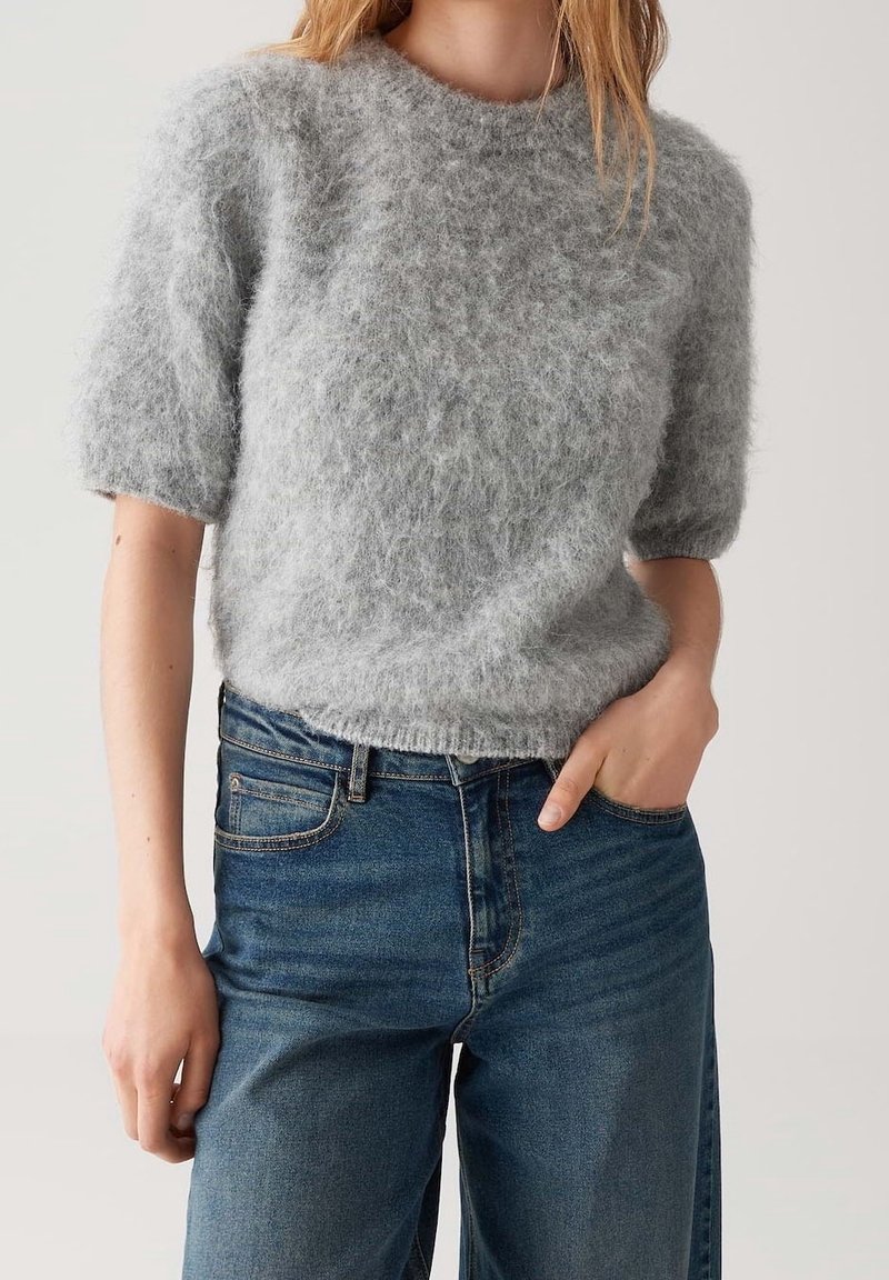 Femme portant un pull court à manches courtes gris clair et duveteux rentré dans un jean bleu taille haute, une main dans la poche.