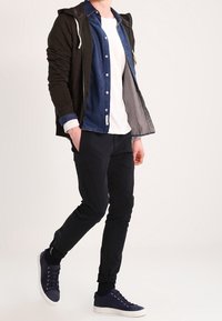 Musta huppari naruilla sinisen farkkupaidan, valkoisen T-paidan, tummien housujen ja laivastonsinisten lenkkarien päällä. Ominaisuuksina on kerroksellinen ilme ja rento tyyli.