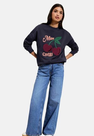 Woman wearing navy sweatshirt with large cherry graphic and "Mon Chéri" text, paired with wide-leg blue jeans and white shoes.