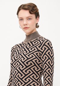 Strikkegenser med høy krage, geometrisk mønster i beige og svart, med ribbestrikket hals og glatt tekstur.