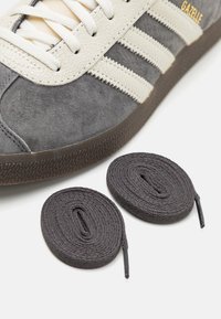 Sapatilhas de camurça cinza com riscas de pele creme e atacadores a condizer. Sola de borracha texturizada. Inclui dois atacadores adicionais em cinza escuro.