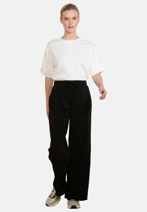 Kobieta idzie, ubrana w białą koszulę z krótkim rękawem, czarne szerokie spodnie ze sznurkiem i beżowe sneakersy na jednolitym tle.