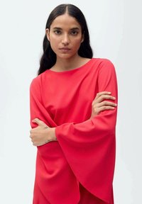 Crvena haljina sa širokim, lepršavim rukavima; glatka tkanina; čamac izrez; model stoji, saša ruke prekrižene, pokazujući oblik i boju odjevnog predmeta.