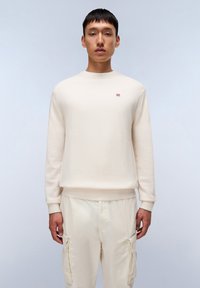 Sudadera color crema con cuello redondo, puños y dobladillo acanalados. Presenta un pequeño logo rojo y blanco en el pecho. Se lleva con pantalones claros.
