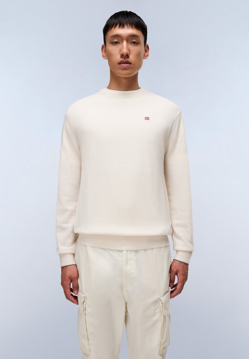 Sudadera color crema con cuello redondo, puños y dobladillo acanalados. Presenta un pequeño logo rojo y blanco en el pecho. Se lleva con pantalones claros.