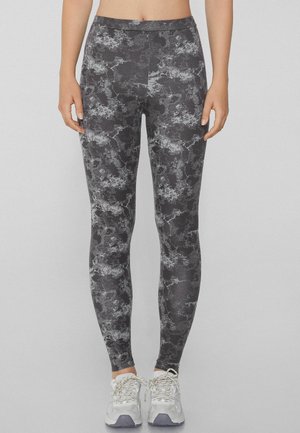 Leggings negros con un patrón abstracto gris. Fabricados con un material elástico, presentan una cintura alta y un ajuste de longitud completa, combinados con zapatillas blancas.