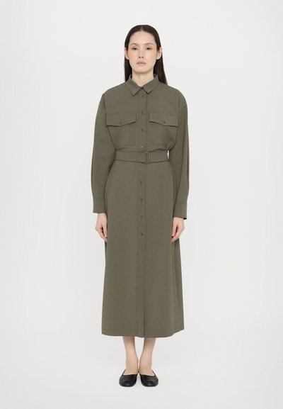 Robe chemise verte olive à manches longues avec deux poches poitrine et une taille ceinturée. Devant boutonné et ourlet à la cheville. Chaussures plates noires.
