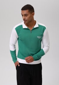 Pullover de media cremallera en verde y blanco, hecho de una tela texturizada. Presenta un diseño en contraste con puños acanalados y el logotipo bordado "ENDURANCE".