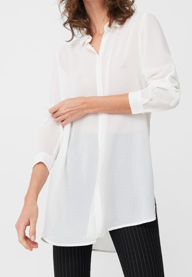 Femme ajustant le poignet d'une longue chemise blanche semi-transparente, portée par-dessus un pantalon noir à rayures.