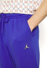 Lila sweatpants med elastisk midjeresår och dragsko, med en svart och vit Jumpman-logotyp på vänster lår. Slät textur.