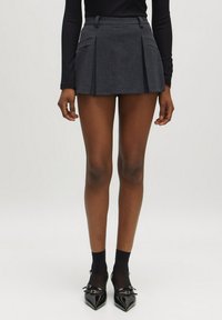 Grijze geplooide mini-jurk met een aansluitende taille, getextureerde stof en twee voorzakken, gecombineerd met zwarte panty en schoenen met een spitse neus.