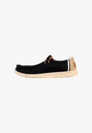Scarpa casual nera senza lacci con suola e tacco beige, caratterizzata da un dettaglio di lacci e cuciture bianche intrecciate sul lato vicino al tacco.