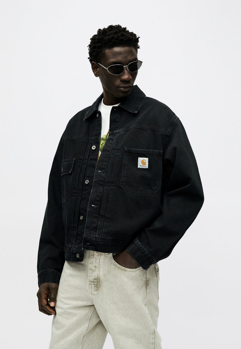 Homme portant une veste en denim noir boutonnée, un jean beige clair et des lunettes de soleil ovales noires, regardant vers le bas à sa gauche avec la main dans la poche.