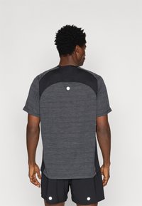 Αθλητικό γκρι κοντομάνικο πουκάμισο με υφασμάτινες πλάγιες επιφάνειες, που διαθέτει αντανακλώσες λεπτομέρειες και ελαφρώς καμπυλωτό στρίφωμα. Φοράει μαύρες βερμούδες.