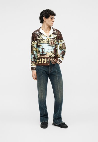 Νεαρός με κατσαρά μαλλιά φοράει πουκάμισο με σχέδια μακρυμάνικο, μπλε τζιν με ραφές μπροστά, μαύρα παπούτσια, γυαλιά και χρυσή αλυσίδα στον λαιμό, όρθιος.