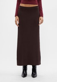 Kobieta w ciemnobrązowej prążkowanej spódnicy maxi, bordowej bluzce z długim rękawem i czarnych błyszczących botkach na obcasie, stojąca na białym tle.