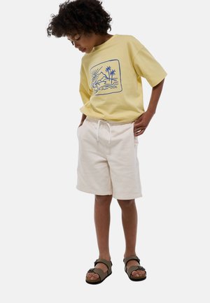 T-shirt en coton jaune avec un graphique bleu représentant un paysage, associé à un short beige avec une taille élastique, porté avec des sandales marron.